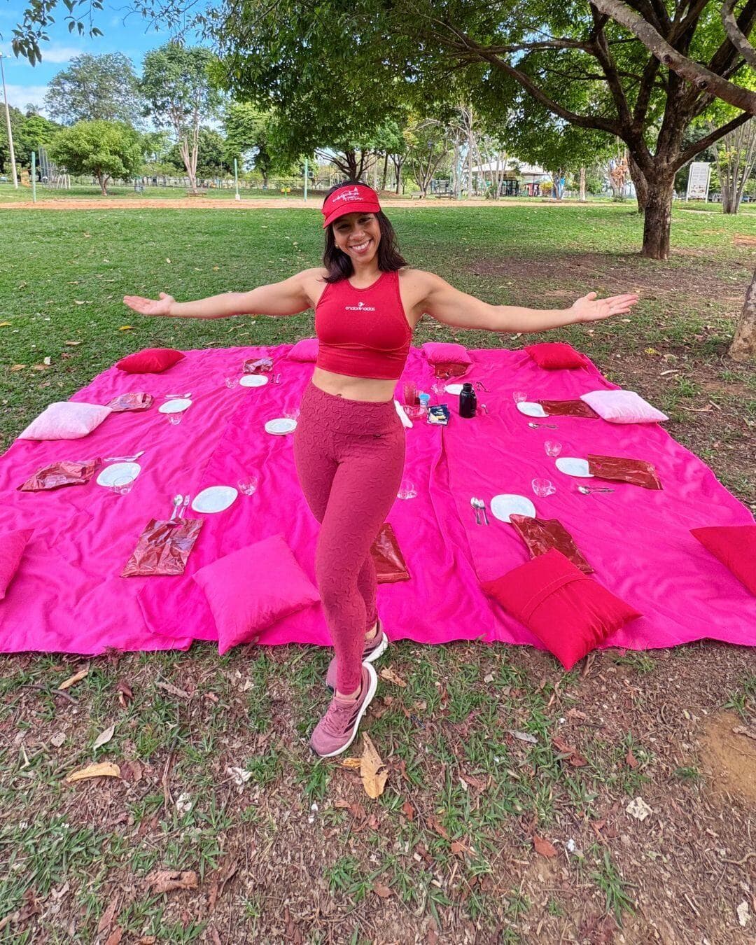 Fernanda de braços abertos sobre a toalha de piquenique rosa, com a mesa posta no gramado do parque