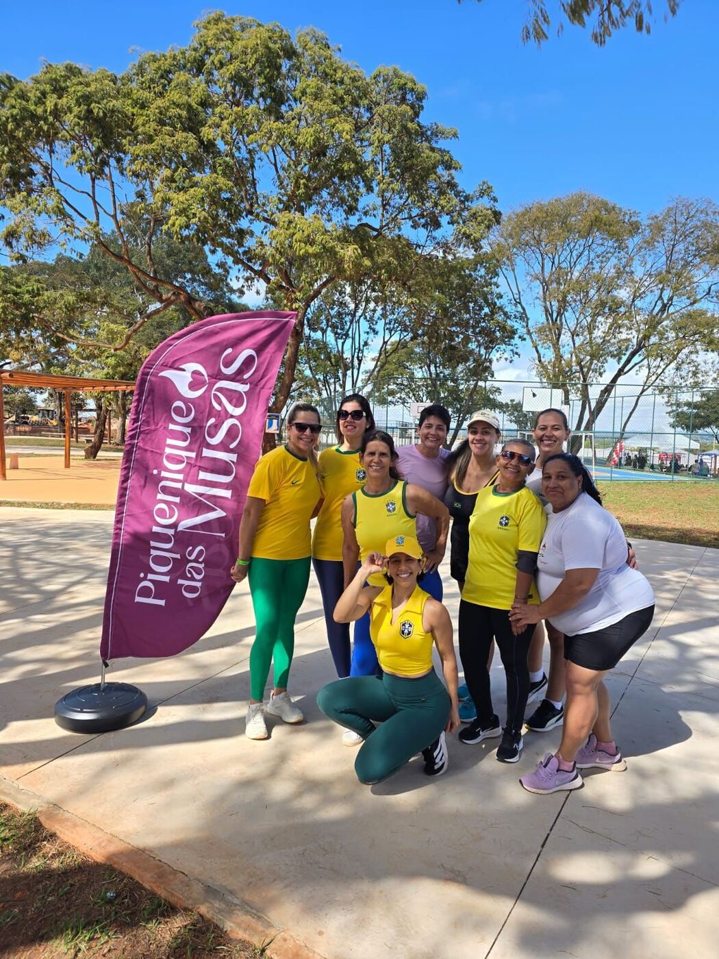 Grupo de mulheres posando ao lado da bandeira do Piquenique das Musas, no parque, em um dia de céu azul
